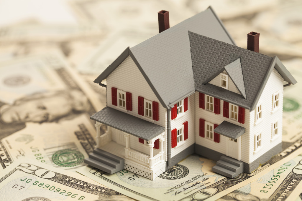 A close-up view of a miniature house placed on top of laid-out twenty-dollar bills, signifying the housing market
