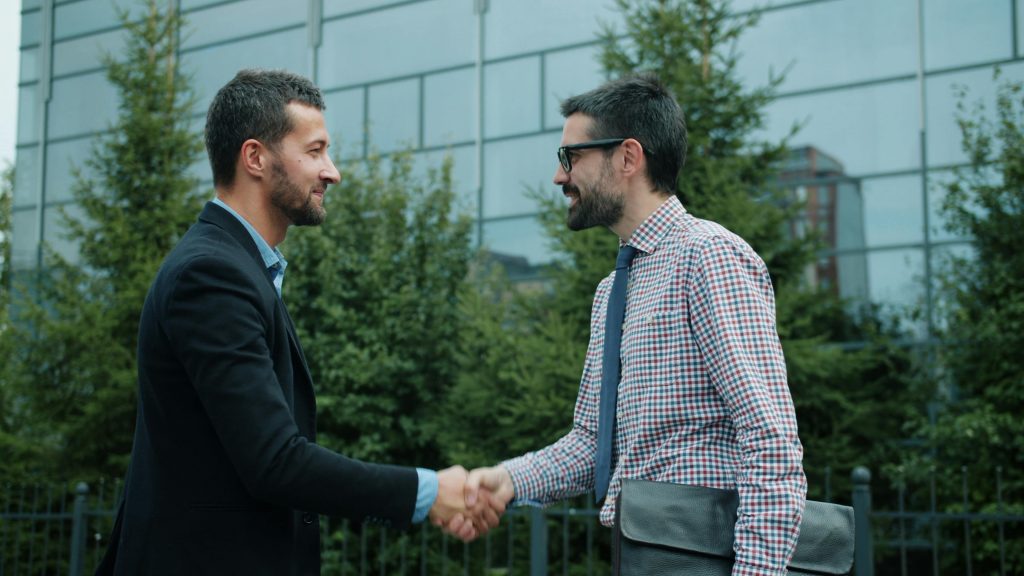 two professionally dressed men shaking hands in front of a commercial building